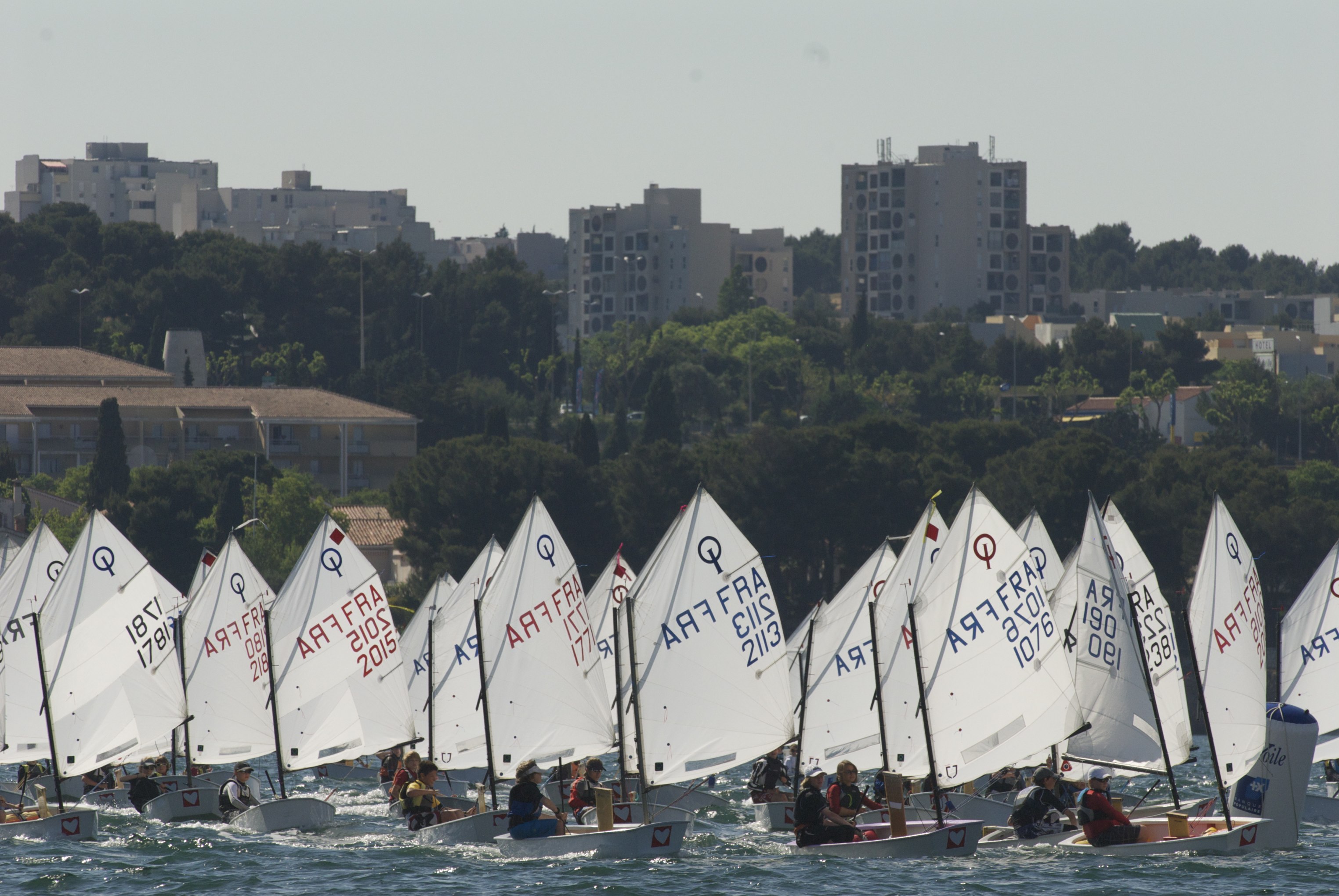 Optimist sailing fleet with white sails along the Mediterranean coast; youth sailing school